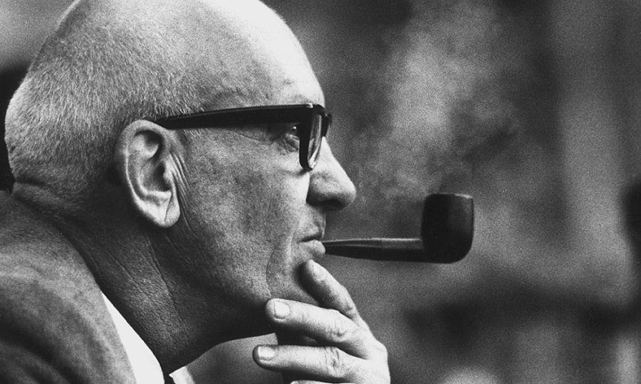 A profile of Mr. Kauffman wearing thick-rimmed glasses and smoking a pipe.