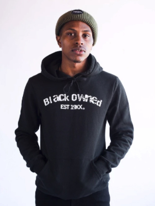 Black model wearing a green knit hat poses in a black hoodie with the message, "Black Owned, Established 19XX," across the front in white lettering.