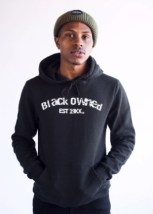 Black model wearing a green knit hat poses in a black hoodie with the message, "Black Owned, Established 19XX," across the front in white lettering.