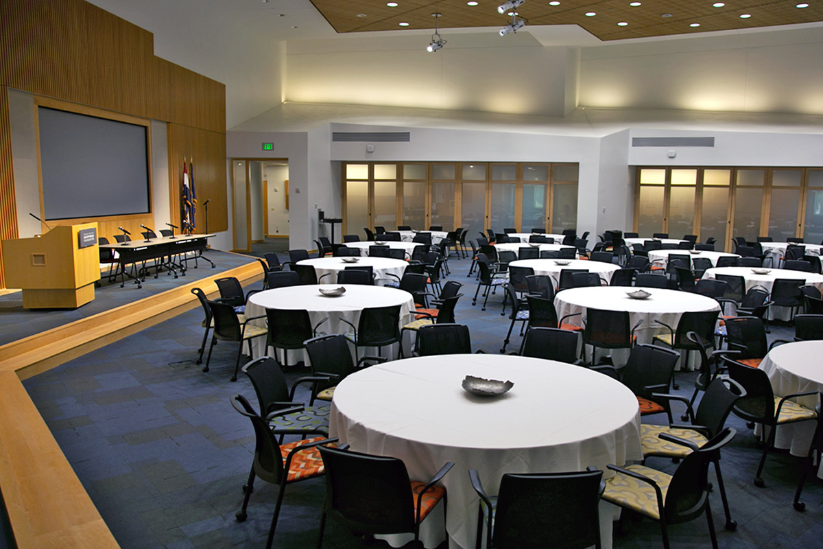 A large conference room featuring circular, cloth-covered tables which seat 200 people, and a stage with podium and another panel table at the front.