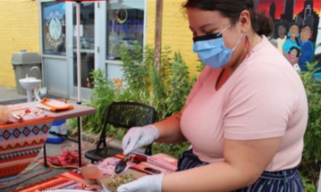 A person wearing a mask and gloves scoops food into a to-go container at an outdoor farmers market