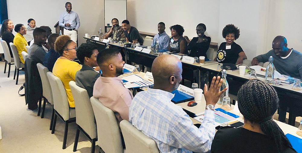 Nineteen people sit together in conversation at two long conference room tables at a Builders and Benefactors convening