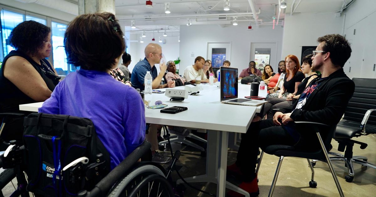 A cohort of sixteen entrepreneurs with disabilities part of the 2Gether-International accelerator program sit around a long table inside a coworking space.