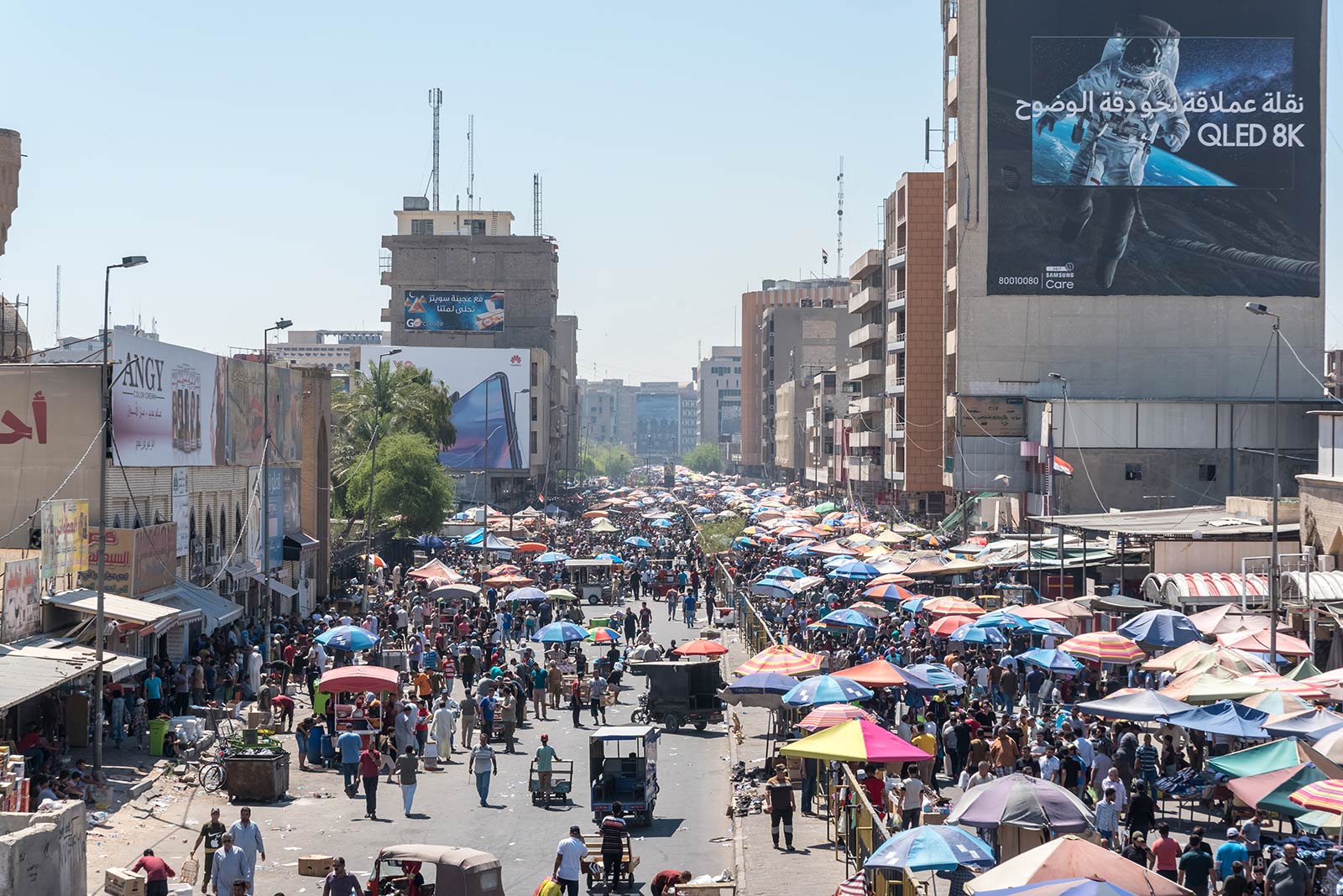 A wide-angle photo of Baghdad, Iraq, shows dozens of people walking down a city street holding umbrellas to block the sun. A building in the background features a large banner image of an astronaut.