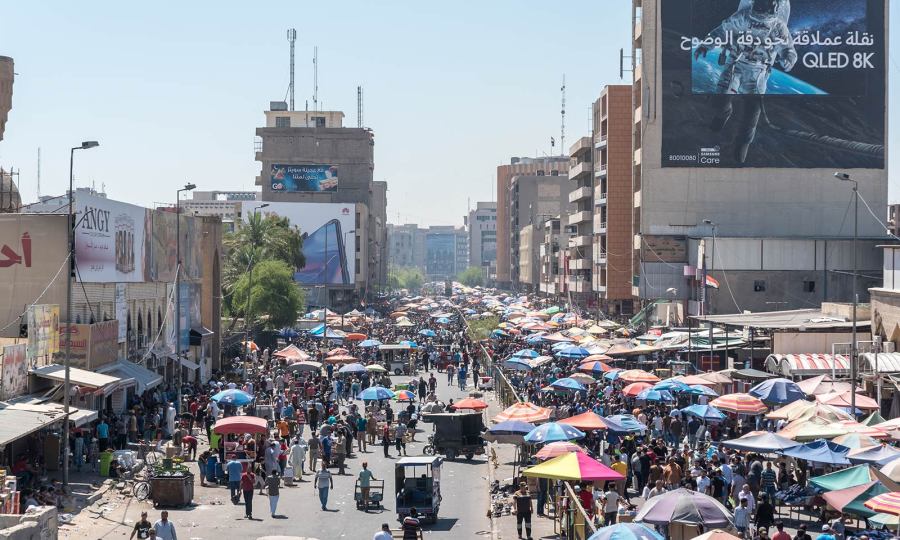 A wide-angle photo of Baghdad, Iraq, shows dozens of people walking down a city street holding umbrellas to block the sun. A building in the background features a large banner image of an astronaut.
