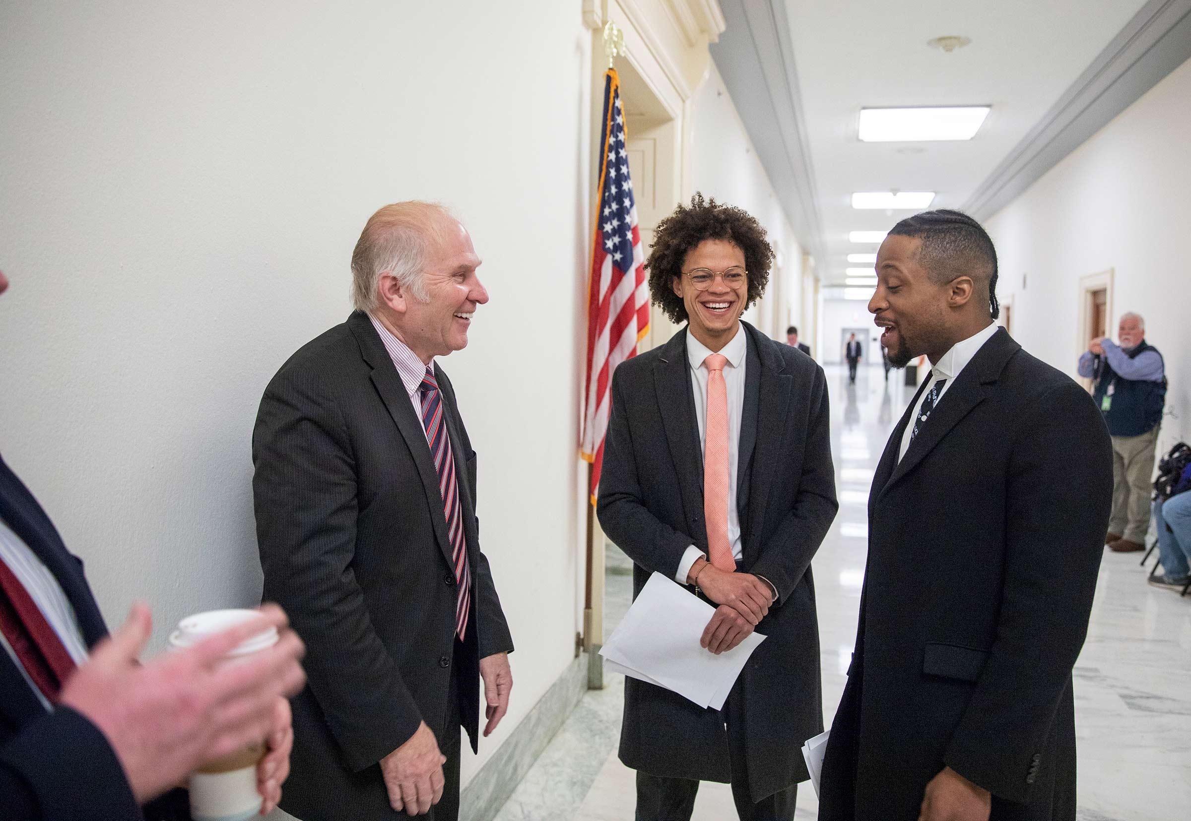 Two entrepreneurs of color wearing suits and ties talk with a white politician, also wearing a suit and tie, in a brightly-lit hallway, an American flag hanging behind them.