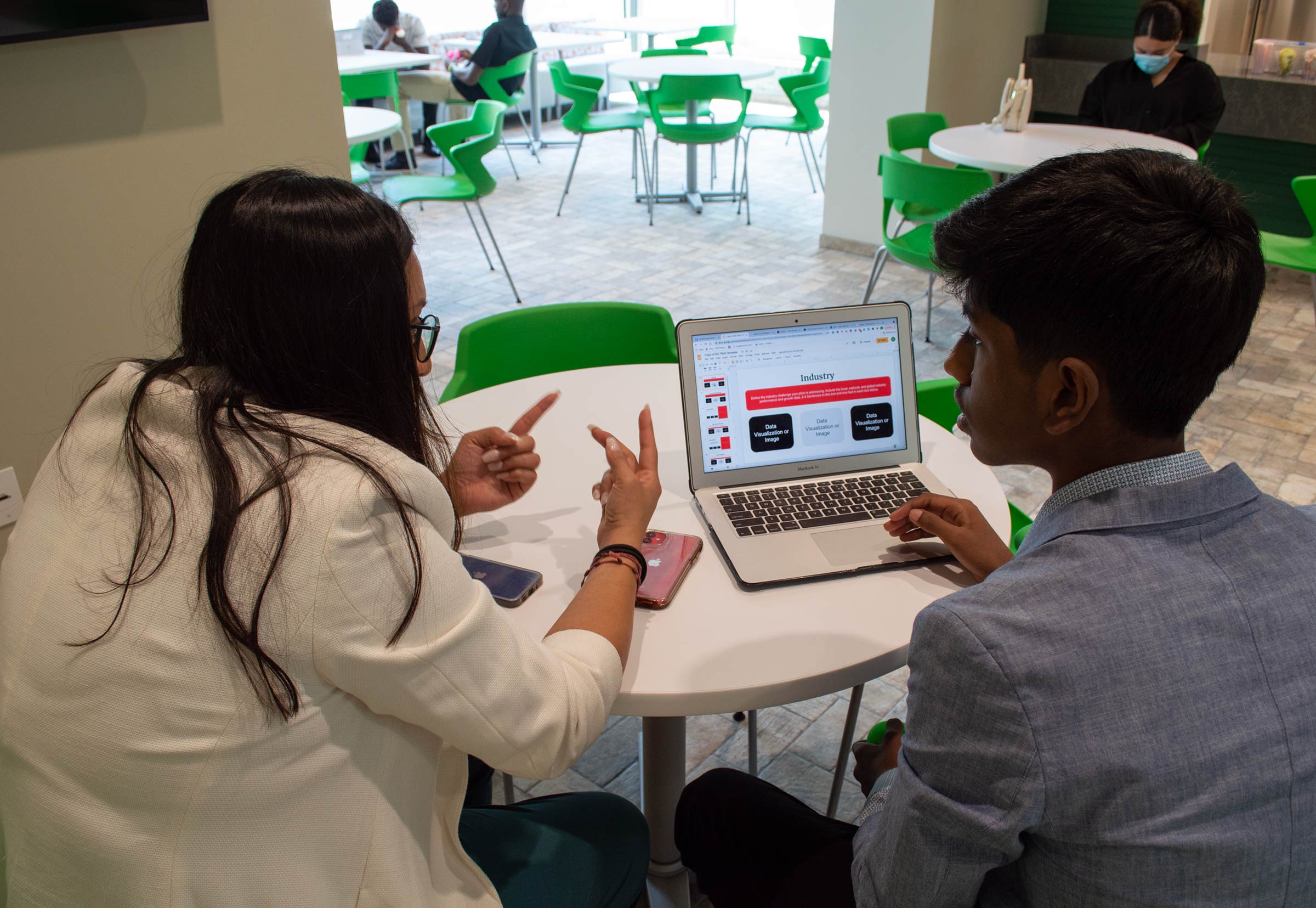 Two people sit at a small round table with a laptop and two phones between them.