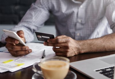 A person holds a smartphone in one hand, a credit card in the other. On the table in front of them are financial documents, a cup of coffee, and a laptop computer.