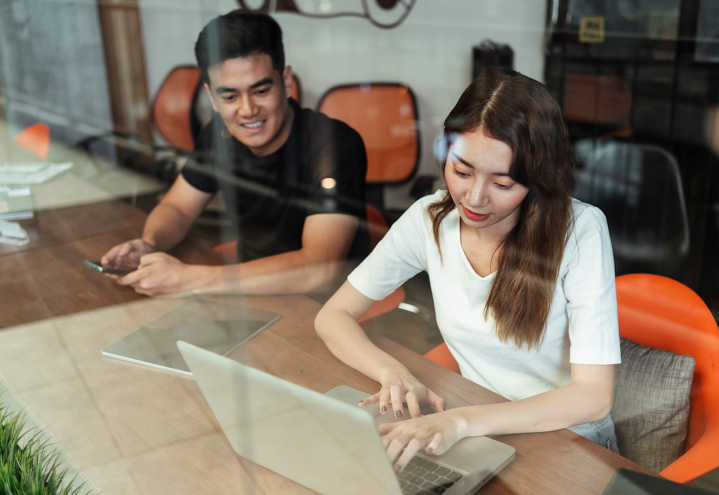 Two people sit in orange chairs at a table, working together on a laptop computer.