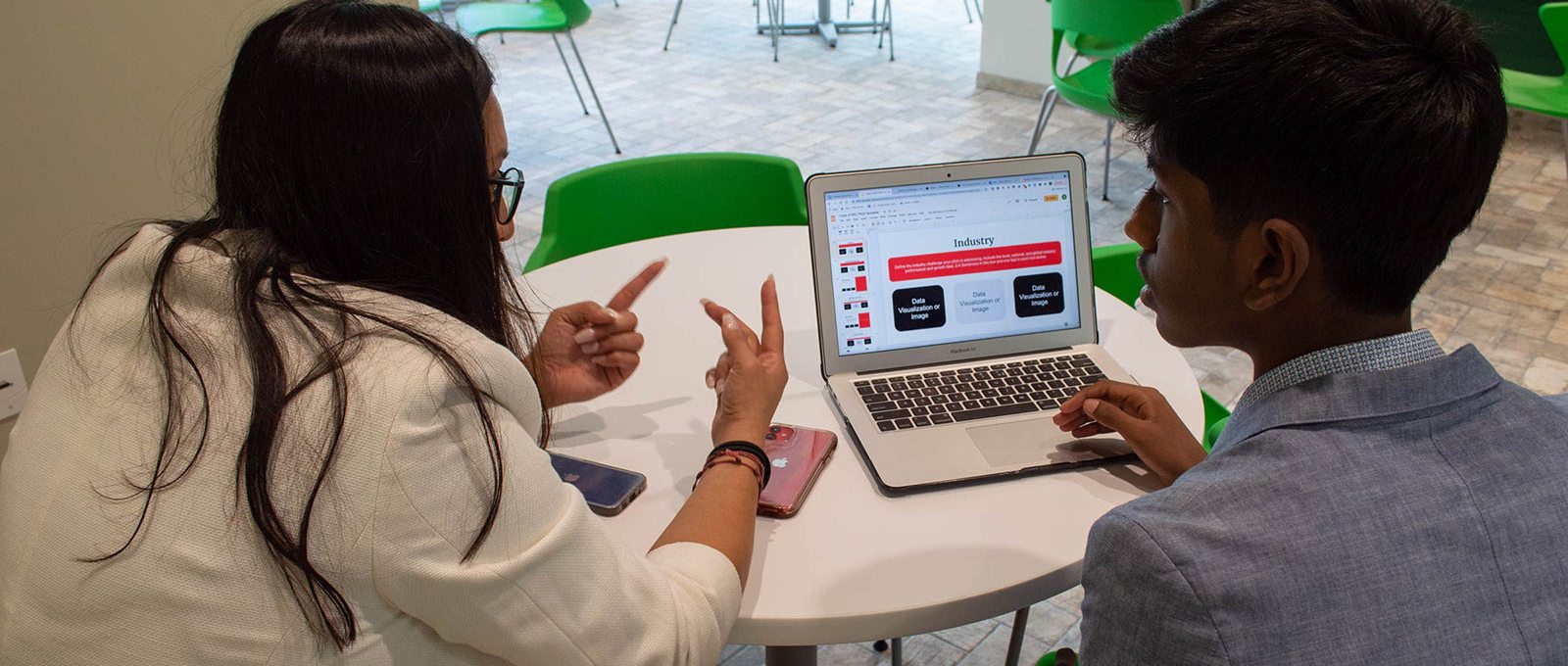 Two people sit at a small round table with a laptop and two phones between them.