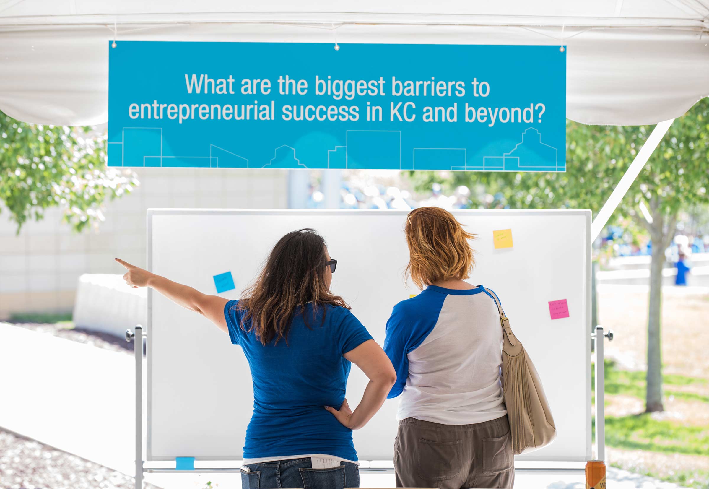 Two people stand underneath a tent outside. A blue banner hanging reads, "What are the biggest barriers to entrepreneurial success in KC and beyond?"