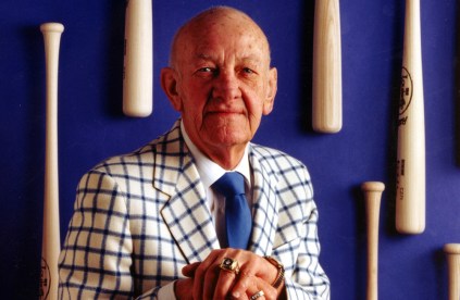 Ewing Kauffman sits in front of a royal blue wall with baseball bats attached to it. He wears a white and blue checkered suit.