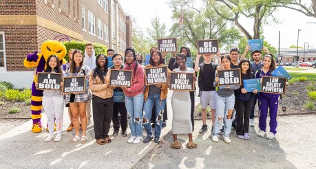 A group of high school students stand outside holding signs which read (from left to right): "I am bold", "Be KC Scholars", "Boldly changing the world", "I am boldly powerful", and "Be bold".