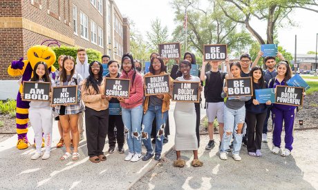 A group of high school students stand outside holding signs which read (from left to right): "I am bold", "Be KC Scholars", "Boldly changing the world", "I am boldly powerful", and "Be bold".