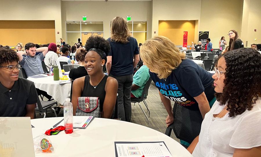 Three students with varying racial and ethnic backgrounds sit at a round table inside a conference room with hundreds of other peers.