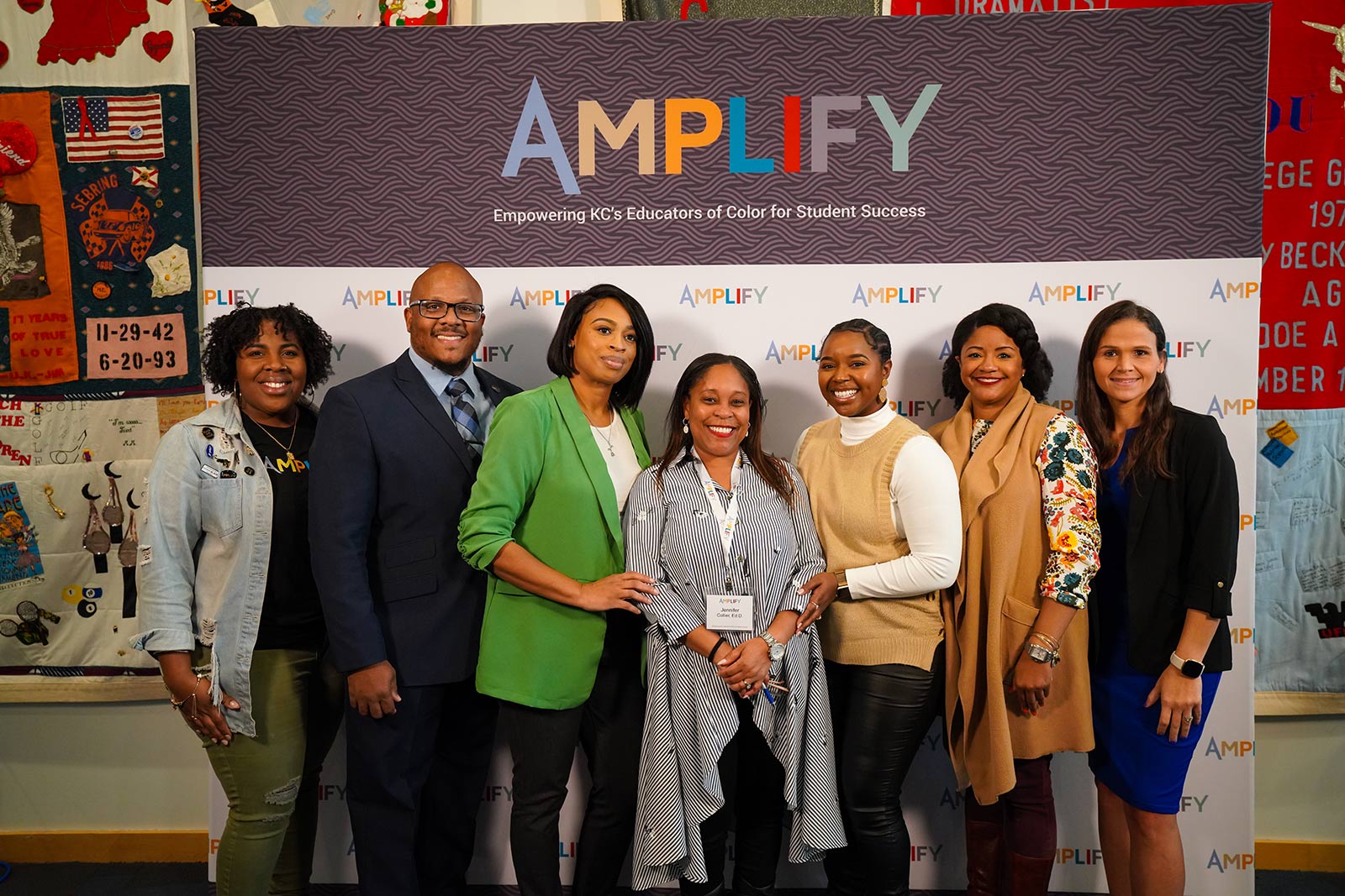 Seven people pose for a photo in front of a backdrop that says "Amplify" in multiple colors. 
