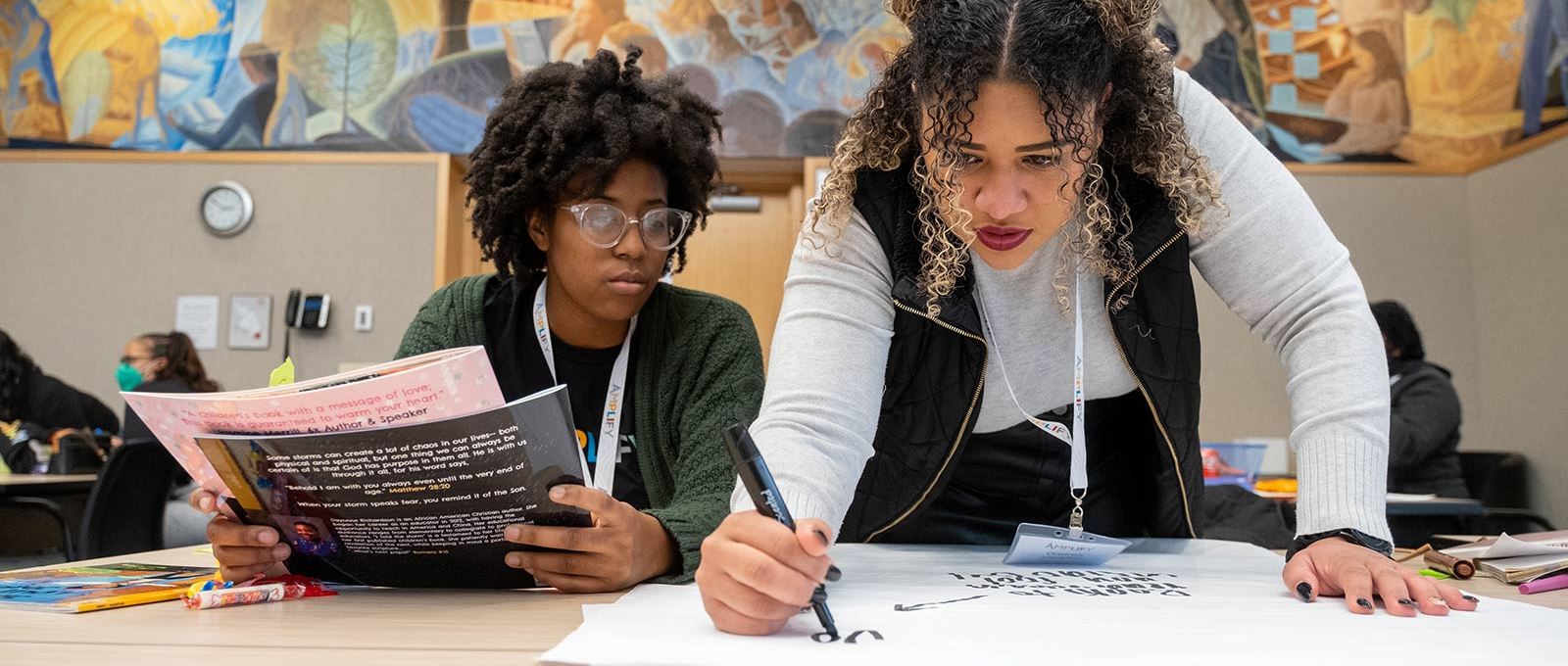 Two racially diverse Amplify attendees sit at a table, one holding pamphlets and the other taking notes.