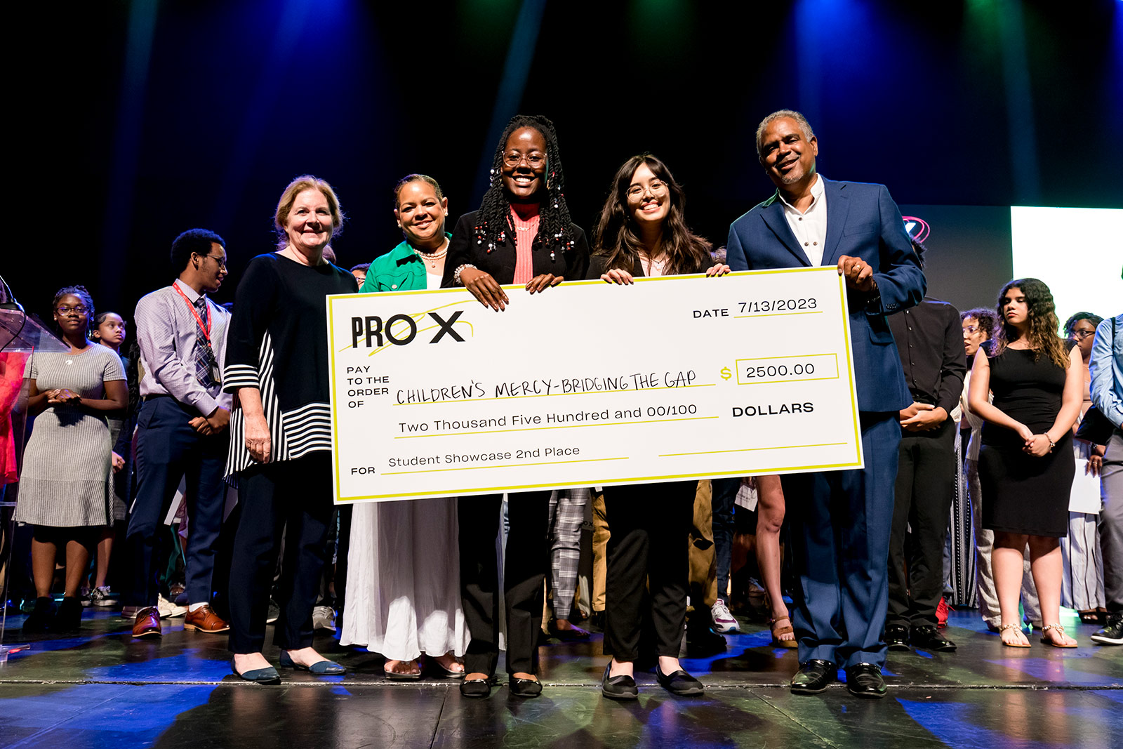 ProX students stand on stage holding a life-sized check made out to Children's Mercy – Bridging the Gap for $2500.