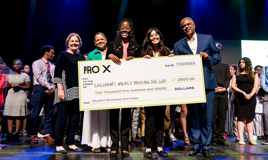 ProX students stand on stage holding a life-sized check made out to Children's Mercy – Bridging the Gap for $2500.