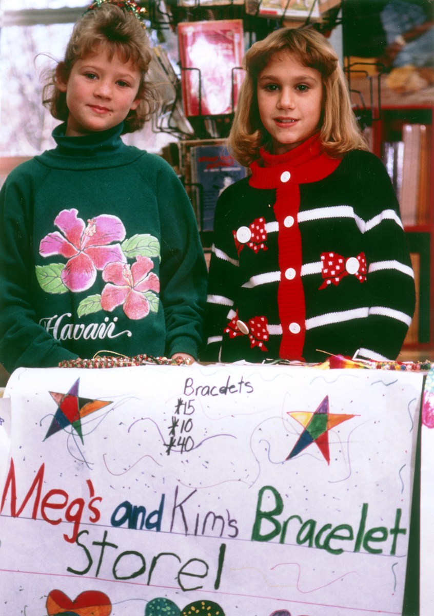 Two young girls stand in front of a table full of bracelets and a decorated sign that says, "Meg's and Kim's Bracelet Store!" while taking part in The New Youth Entrepreneur (YESS!) program.